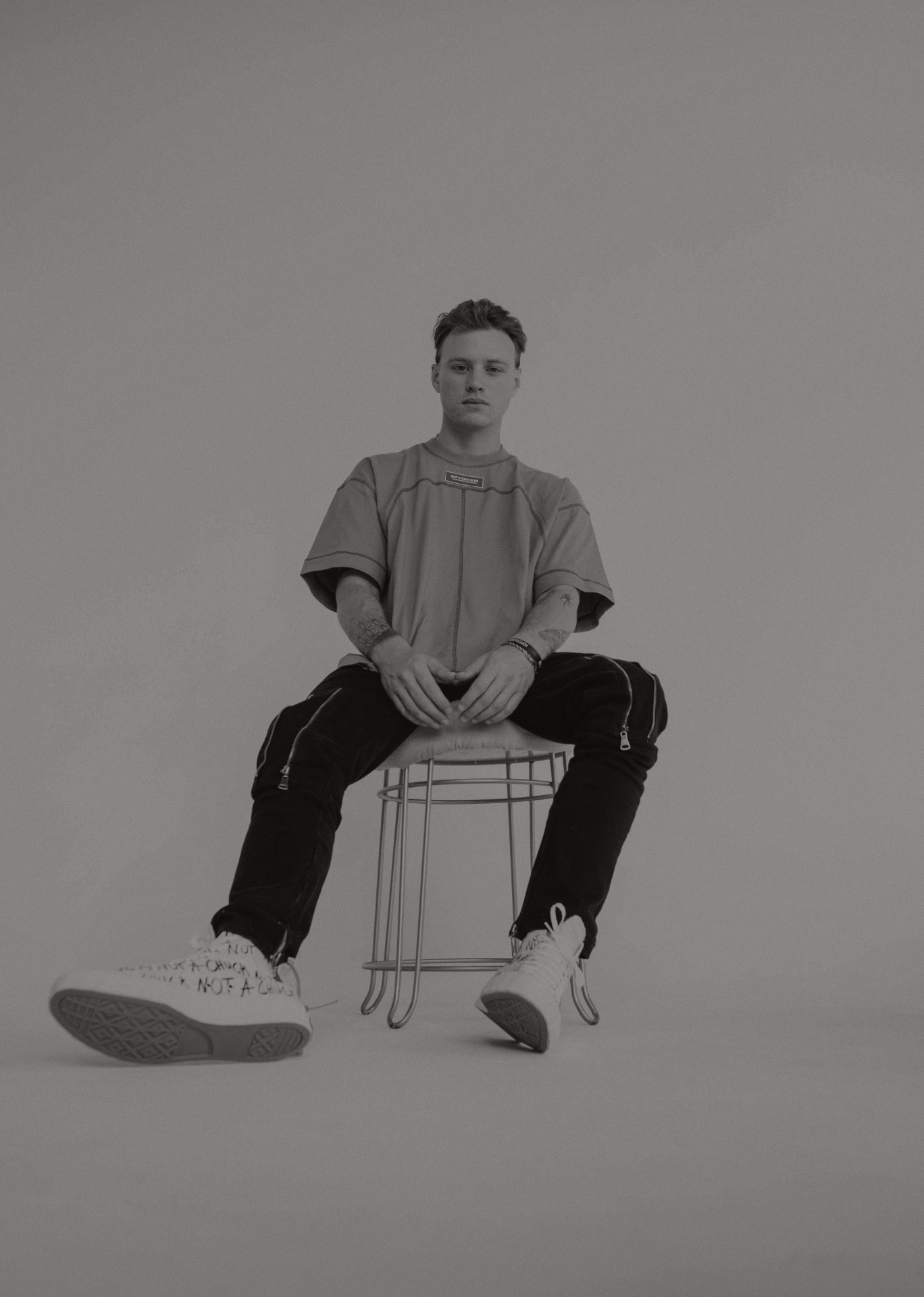 OPTIMISM. archive — Contrast-stitch tee and zip trouser. Seated study, single stool.